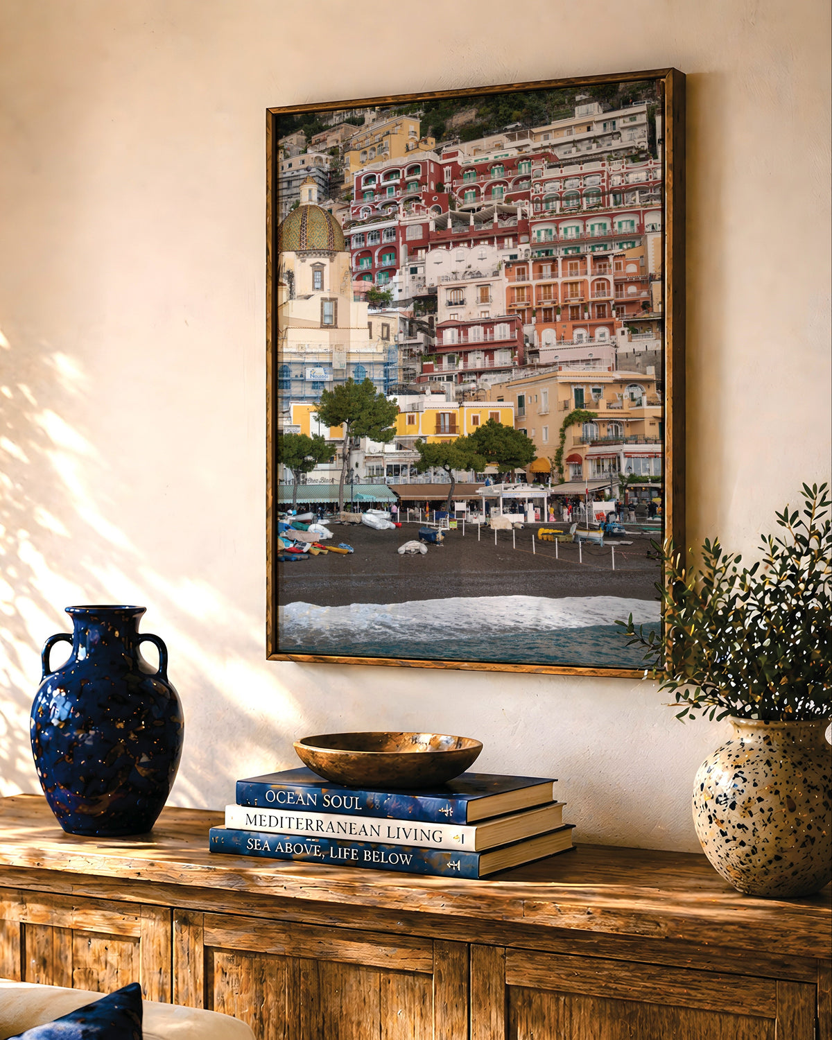 A framed coastal art print depicting colorful Mediterranean hillside buildings and a beach is displayed on a beige wall above a rustic wooden console table. The room is styled with a blue ceramic vase, books, and potted plants, with warm sunlight casting shadows across the wall.