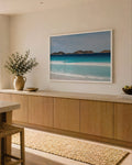 Framed Lucky Bay landscape print of turquoise surf and rocky islands in a white frame, hung above oak cabinetry in a bright coastal kitchen with a jute runner and ocean view.