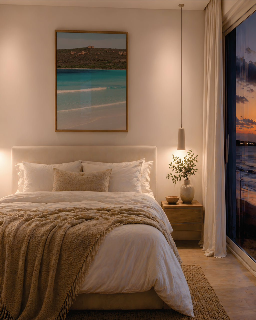 Framed beach print of turquoise water and coastline in an oak frame, above a bed in a warm coastal bedroom at dusk, with floor-to-ceiling windows overlooking a sunset ocean view.