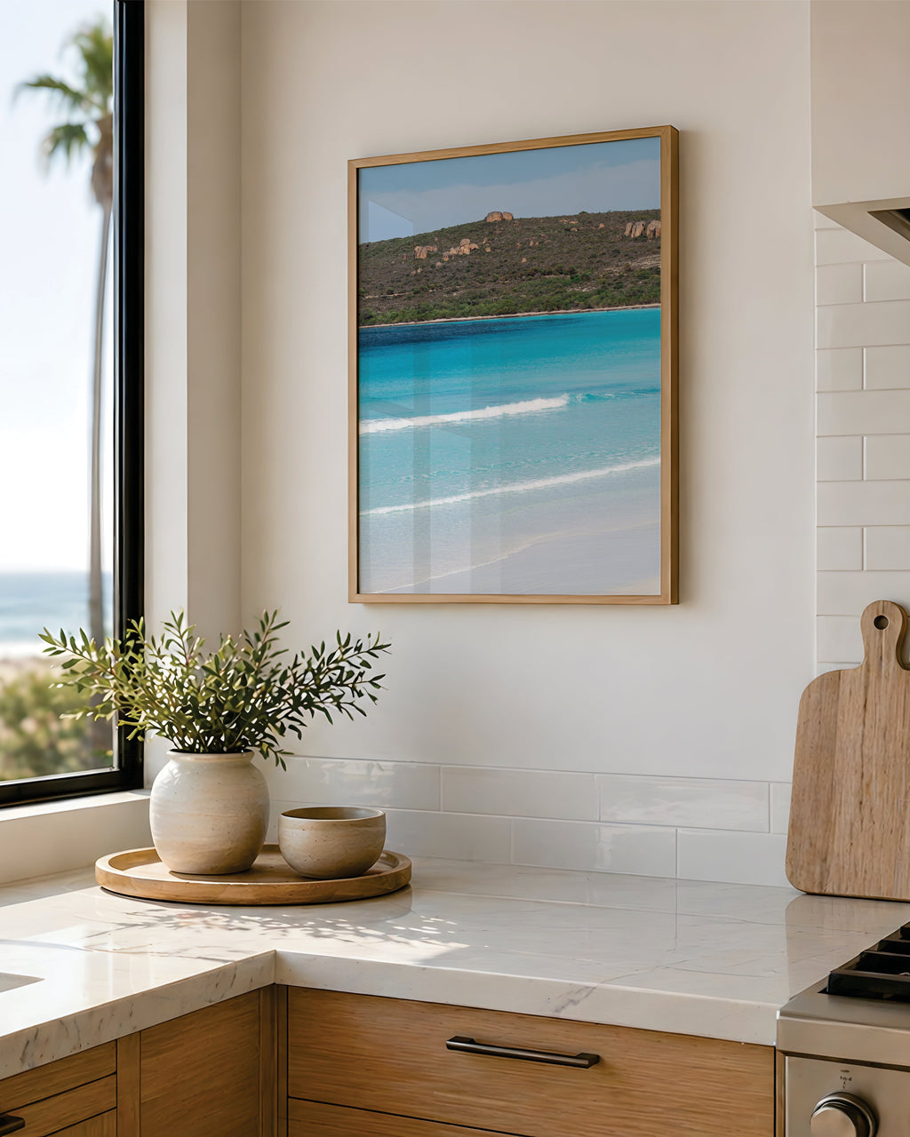 Framed coastal print of a turquoise bay and white sand beach in a light oak frame, hung in a bright modern kitchen with marble benchtops and an ocean view window.