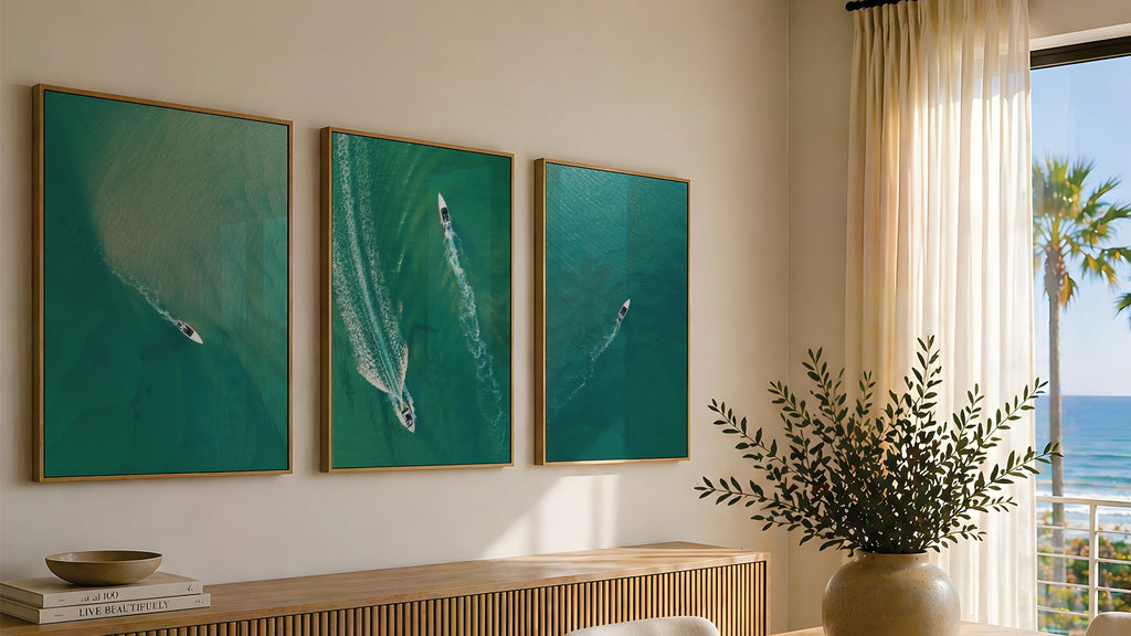 A modern dining room scene with three framed teal ocean art prints hanging above a wooden sideboard and dining table. The room has cream walls, sheer curtains, a large vase with greenery on the table, and a window overlooking palm trees and the sea.