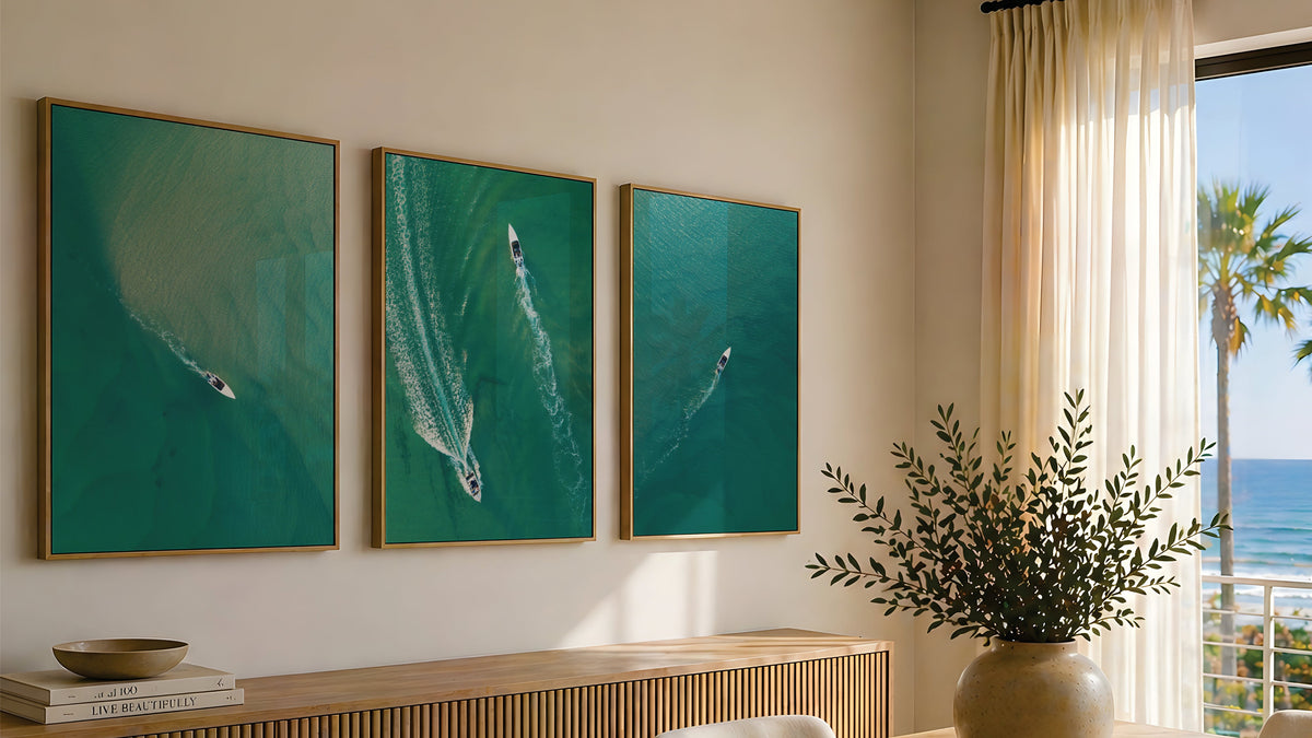 A modern dining room scene with three framed teal ocean art prints hanging above a wooden sideboard and dining table. The room has cream walls, sheer curtains, a large vase with greenery on the table, and a window overlooking palm trees and the sea.
